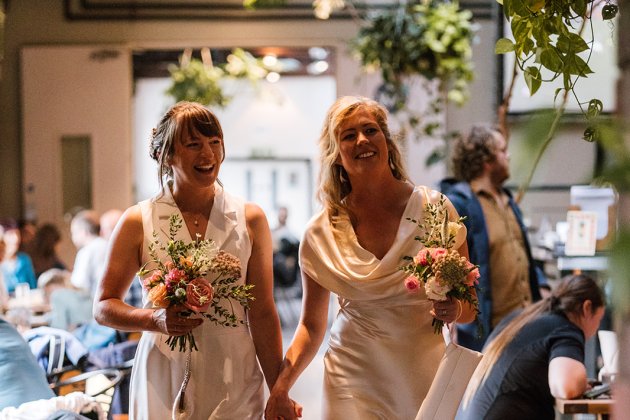 Two brides in white dresses holding bouquets and smiling while walking hand in hand through the Café & Bar at Watershed.