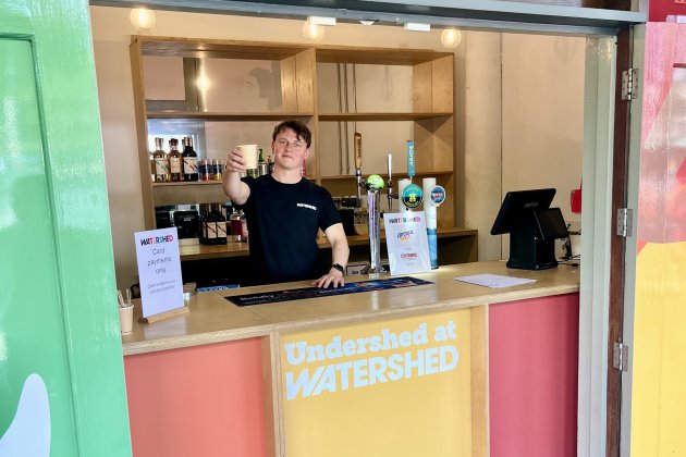 A member of the Café & Bar team holds a drink to the camera from the Undershed Bar.