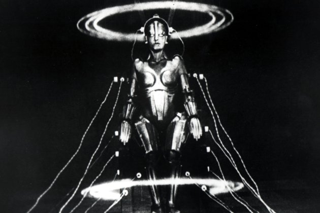 A black and white image of a humanoid robot with wires attached in a darkened room