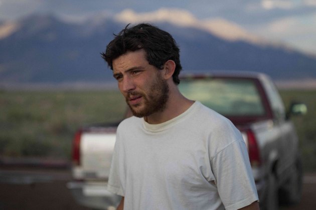 A man wearing a white t-shirt looks off into the distance.