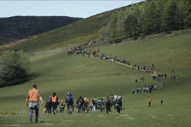 Still from Our Land of a large group of people walking up a grassy hill.