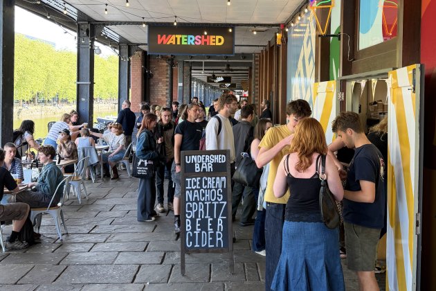 People gathered outside Watershed venue with a chalkboard menu offering ice cream, nachos, spritz, beer, and cider on a sunny day.