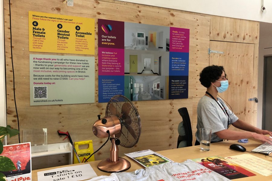 The hoarding outside of Watershed's toilet building works, behind Watershed's welcome desk. A member of staff is at the desk and you can see signage behind them.