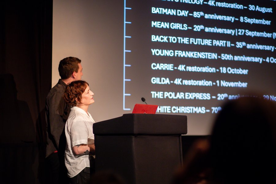 Two people presenting on a stage in front of a cinema screen