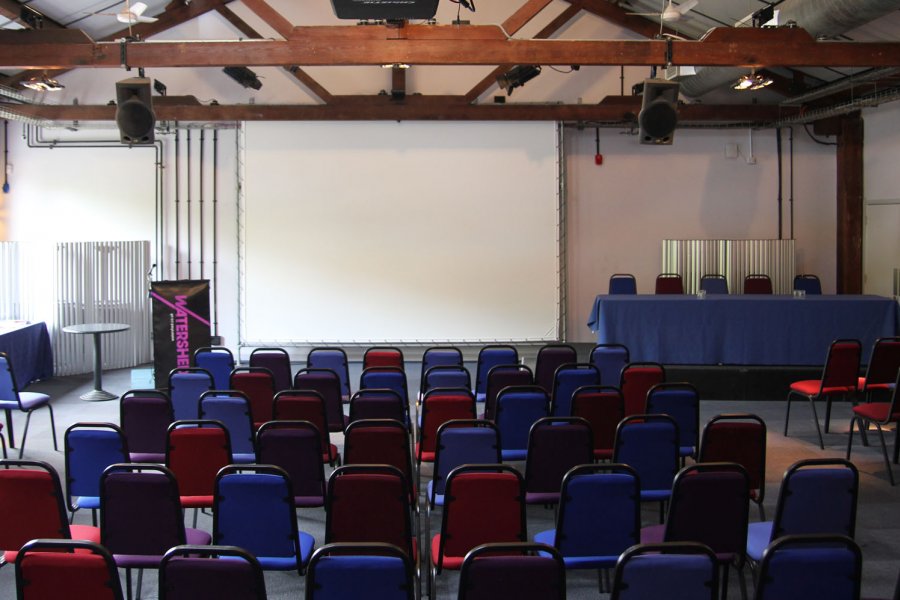 Photo of Waterside 3 at Watershed – a large room with chairs facing a projector screen, a speakers podium and top table