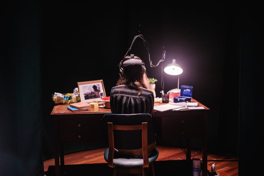 In a dimly lit room a woman sits at a desk wearing a headset. Her back is to us.