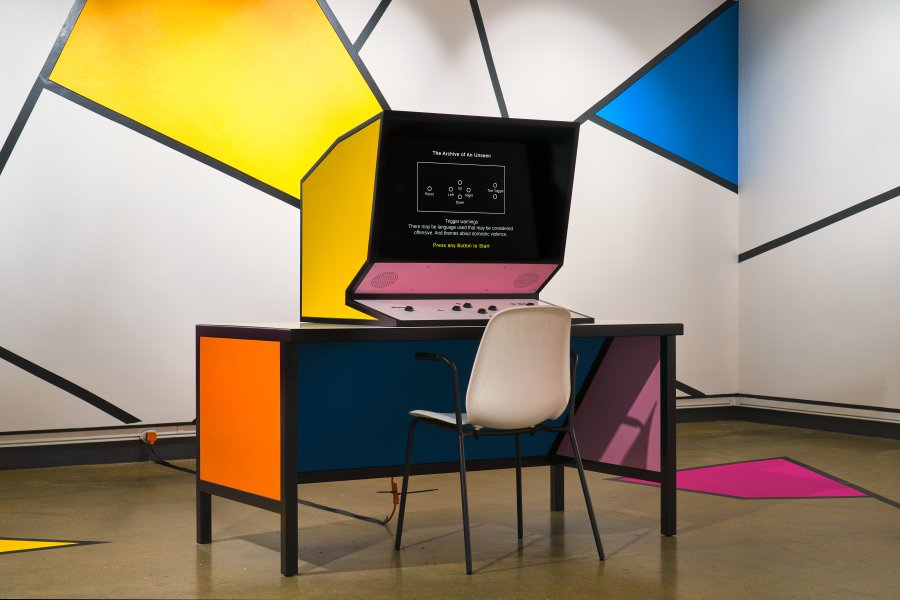 A large, brightly coloured computer sits on a brightly coloured desk. In the background, a wall is painted in bold, bright gemoetric shapes.