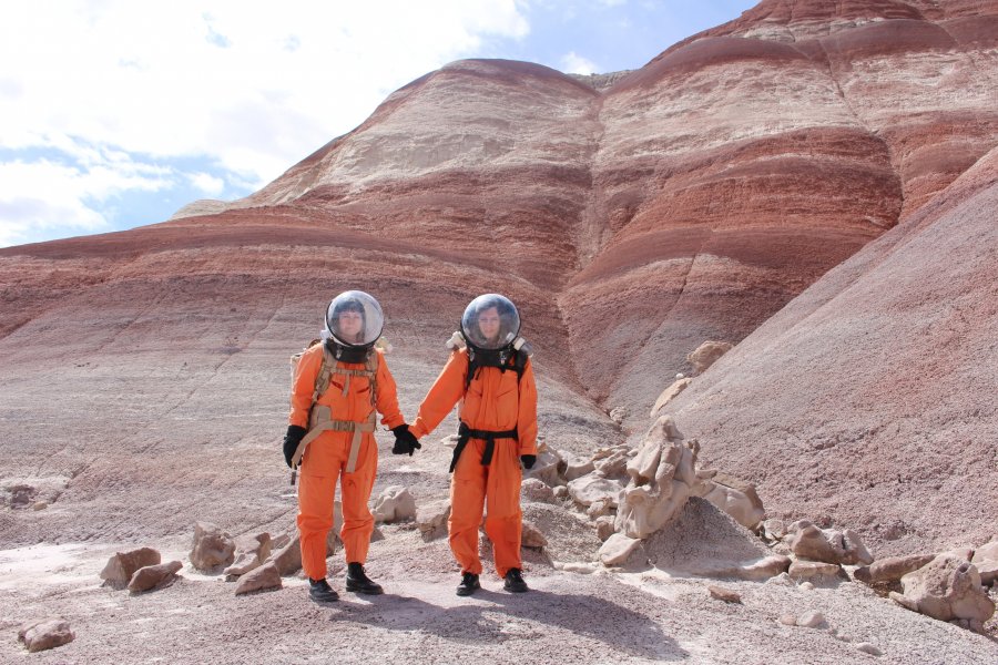 Two women in spacesuits in a barren landscape holding hands