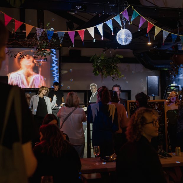 A lively party scene featuring a crowd watching a musician perform on a screen, with colourful decorations and a DJ setup in the Watershed Café & Bar.