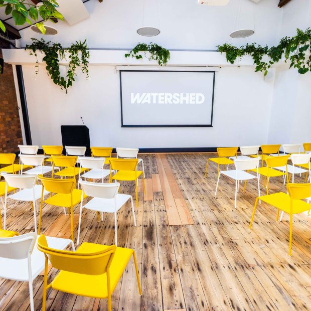 Bright, modern event space with yellow chairs arranged in rows, a projector screen displaying "WATERSHED," and greenery for decor.
