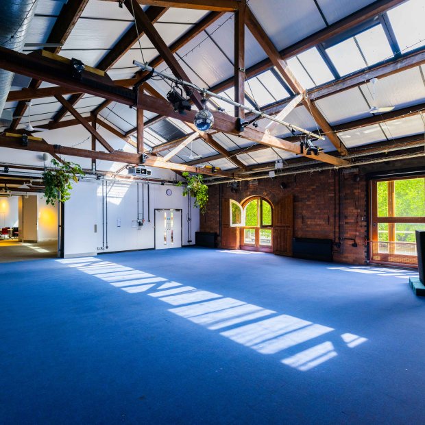 Spacious, sunlit room with high wooden beams, blue carpet, and large windows, featuring greenery and a minimalist podium.