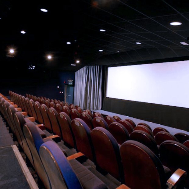 A dimly lit cinema hall featuring rows of red upholstered seats facing a large white screen, with curtains on one side.