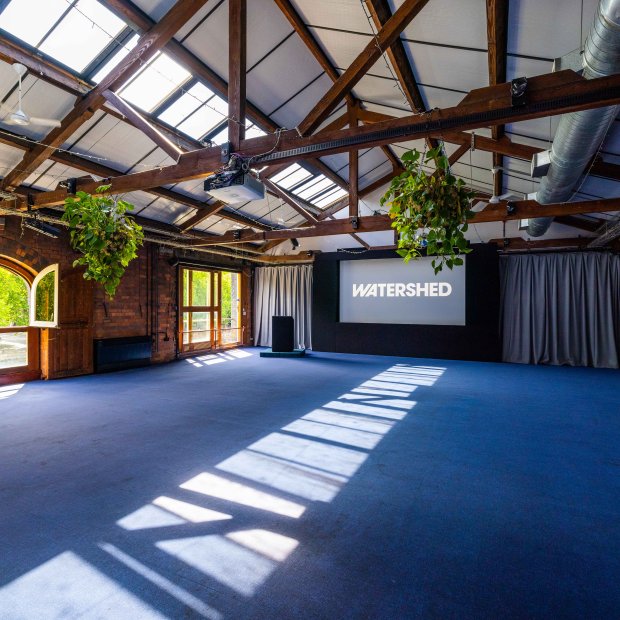 Open heritage event space with exposed timber beams, skylights, hanging plants, and a screen displaying “WATERSHED.”