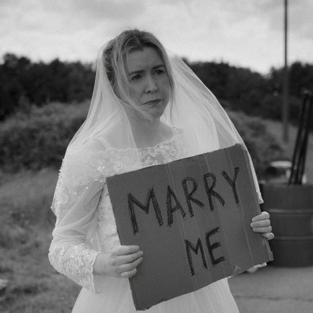 A bride wearing a veil holds a cardboard sign that reads "MARRY ME," standing by a roadside with a mailbox nearby.