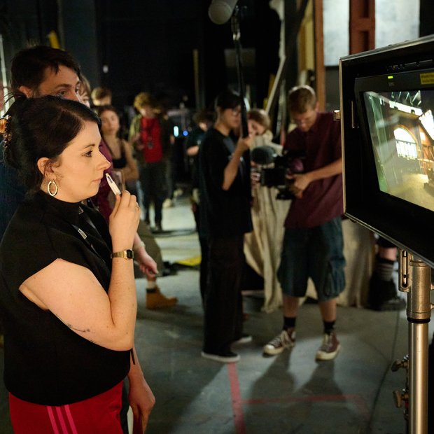 A woman attentively observes a monitor on a busy film set, surrounded by crew members and equipment. The scene conveys focus and collaboration.