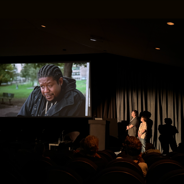 Audience watches a film screening in a dark theater while two presenters stand near a podium beside the screen.