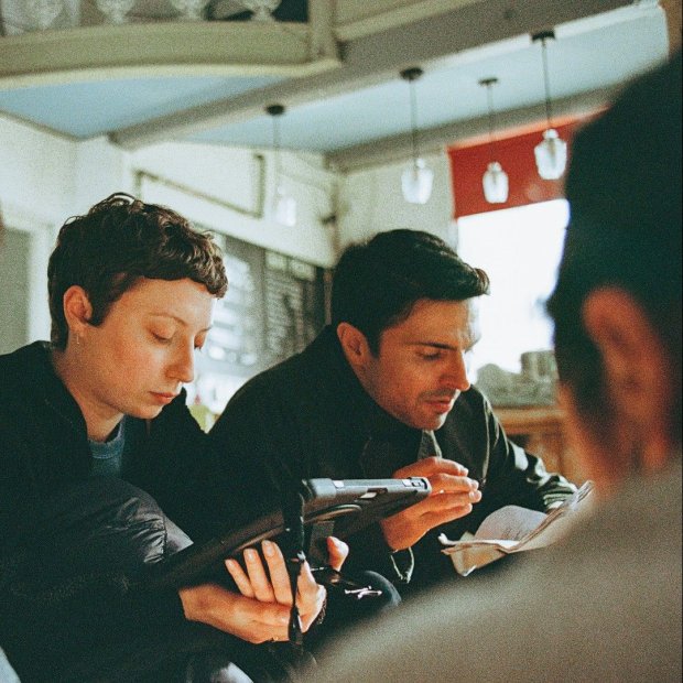Behind the scenes on the shoot of Surviving Earth. Three people sit closely in a cozy café, a women looks at a handheld screen. The background shows warm lighting and a relaxed atmosphere.