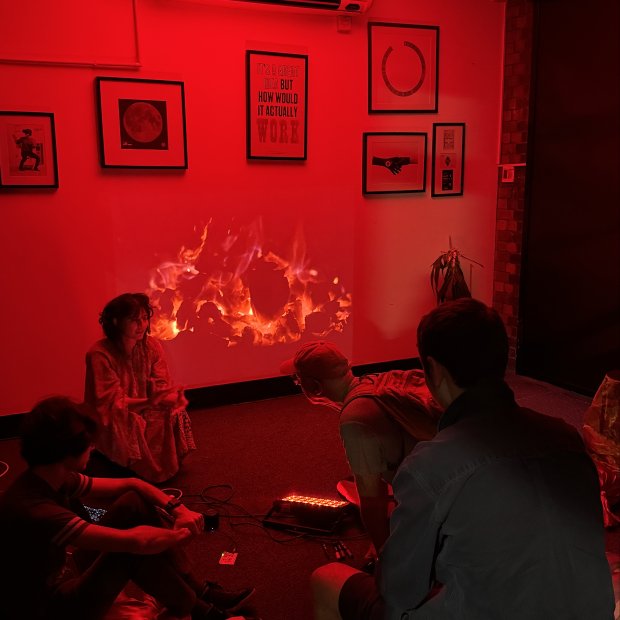 A group of people sit in a dimly lit room with red lighting, focused on a vibrant fire projection on the wall. Modern art hangs above them.