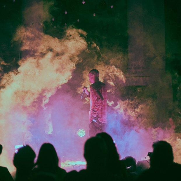 Performer holding a microphone on a foggy, dimly lit stage with an audience silhouette in the foreground.