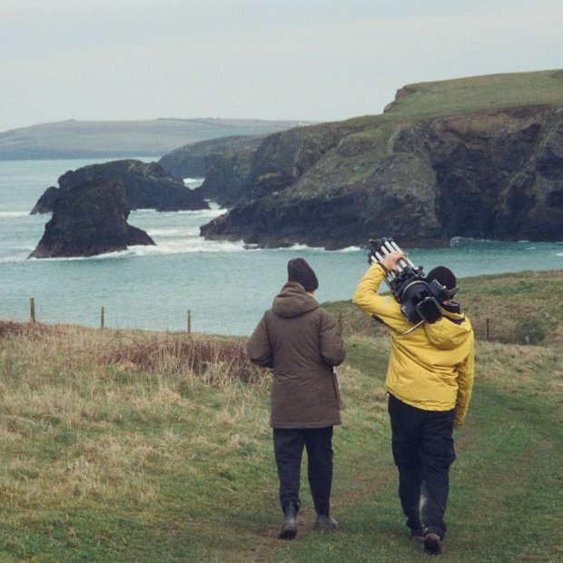 Two people walk along a grassy coastal path with cliffs and ocean in the background, one carrying a camera tripod.