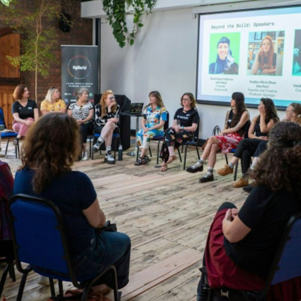 Diverse audience listens to women speakers seated in a circle during a community presentation, with portraits and speaker names projected behind them.