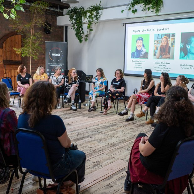 Diverse audience listens to women speakers seated in a circle during a community presentation, with portraits and speaker names projected behind them.