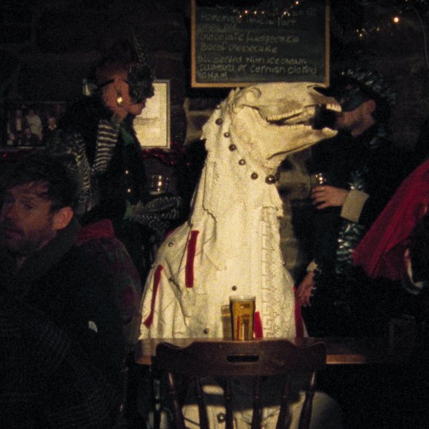 A still from Pen Mari: a white person-sized puppet with a horse's skull perched on the top. Sat amongst a busy, dimly-lit pub.
