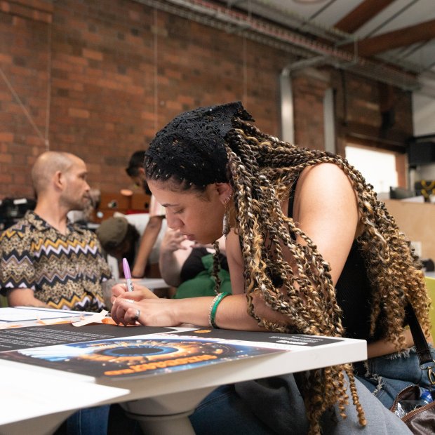 A young woman with long braids leans over a studio table, writing on a large printed sheet as others gather nearby in a lively, creative space.