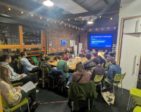 A diverse group of people sit around a rectangular table in a modern meeting room with exposed brick walls. A presentation titled "Key Objectives & Messages" is projected on a screen. The room is well-lit with string lights, creating a collaborative and focused atmosphere.