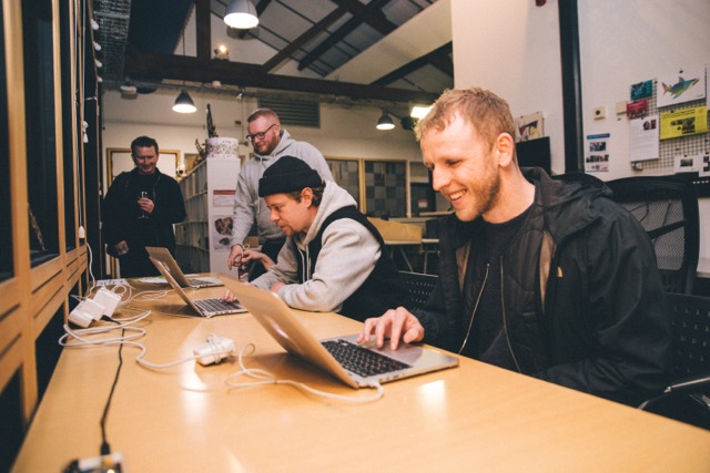 three people sit at laptops, the nearest one smiles at the screen 