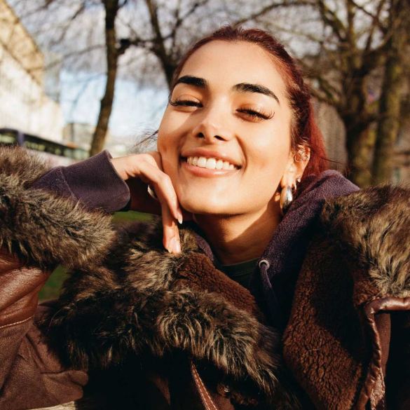 A young olive-skinned woman grins at the screen, she's wearing a brown jacket with a fur collar.