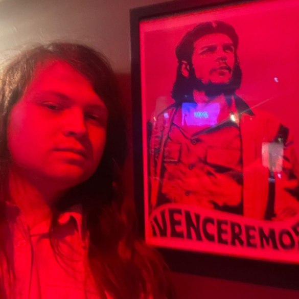 A young white non-binary person stands to the left and there is a red light and a framed photograph next to them.