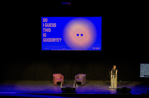 Dark theater stage with a speaker at a lectern, two chairs, and a blue screen reading “SO I GUESS THIS IS GOODBYE?”—reflective tone.