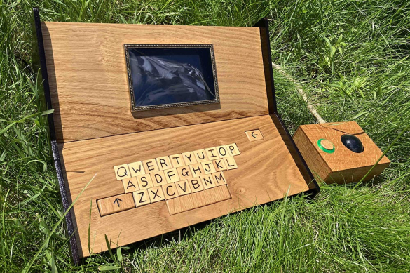 A wooden laptop with a screen and QWERTY keyboard sits on green grass, accompanied by a rectangular control box.