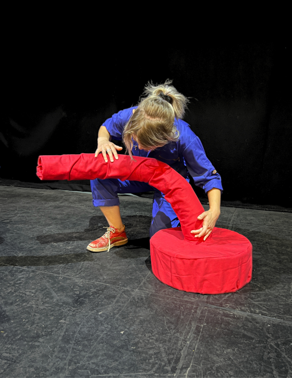 Nat, in  bright blue  overalls interacts with one of their talking beings, which consists of  a red, curved foam piece and a round cushion. The background is a dark stage. 
