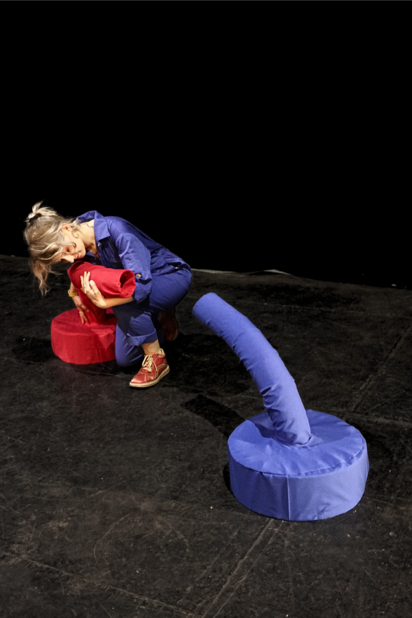 Nat is shown at an angle wearing blue overalls on a dark stage, interacting with their red talking being. Both beings are in the shot facing each other.