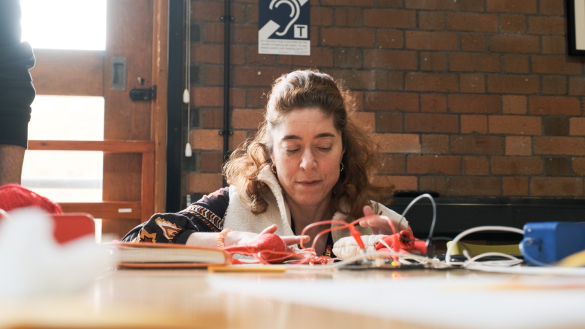 A person sits at a table working with colorful yarn and various electronic components, surrounded by notebooks and tools.