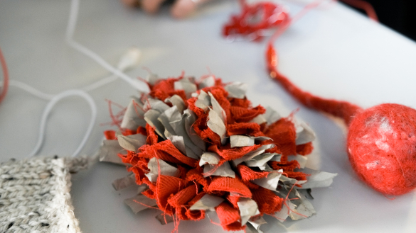 A close-up of a colourful textile flower made of red and gray fabric scraps, with a red felt ball and knitted piece in the background.