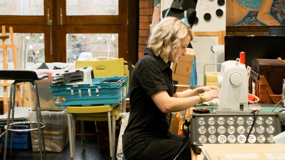 Nat sits at a sewing machine in a cluttered workspace with various tools and materials around them, including chairs and storage boxes.