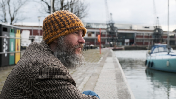 Geraint  sits on a waterfront, gazing into the distance with a hopeful spark in his eye.