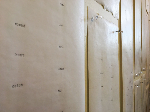 A close-up of typewritten pages pinned to a wall, featuring vertically aligned words and phrases in a minimalist setting.