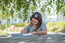 A woman in glasses paints on a table outdoors, under a tree with leaves in the background.