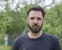 Lukus is a white male with a dark beard and hair. He is wearing a black t-shirt and is standing in front of a number of trees in summer time.