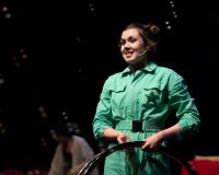 Scarlett stands just right of the centre holding two hula hoops by her waist. She is smiling, surrounded by bubbles and wearing a green boiler suit with a sequin star on the chest pocket. 