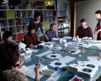 A group of men and women, young and old, seated and standing around a table. They are hand-finishing the Here We Grow books.