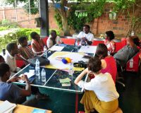 A group of black men and women seated around a table. In the centre of the table are items to make books - paper, string. There are also headphones on the table. Surrounding the people are plants and trees.  