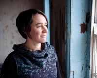 White female with short dark hair looking out of window smiling wearing a patterned blue dress with a large roll neck