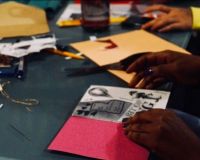 A black woman, seen only by the hands, customises and designs her zine which lays open on a dark green table surrounded by scissors, colourful prints, stamps, pins and other crafting material.