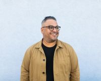 An Image of Nikesh Shukla wearing a yellow shirt with a black t-shirt underneath, he is looking into the distance and smiling.