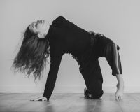 A dancer arches their back with both feet and one hand on the floor. The picture is in black and white. The dancer is wearing dark clothing and has long dark hair. The floor is shiny and wooden
